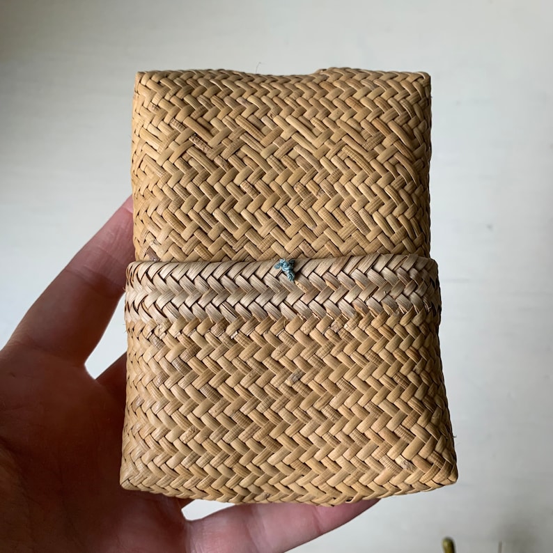 May include: A small, rectangular woven basket with a light brown colour and a woven pattern. The basket has a lid that is closed.