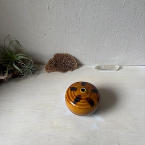 May include: A small, round, glazed ceramic vase with a glossy mustard-yellow finish and dark brown, elongated spots arranged in a circular pattern around a central opening at the top. The vase has a smooth, slightly ribbed texture.