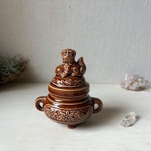 May include: Brown ceramic incense burner with a detailed design. The burner features a decorative lid with a small lion-like figure on top. The base has floral and bird patterns, and small handles. A unique decorative piece.