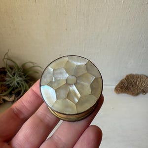 May include: A small, round, lidded container featuring a geometric mosaic pattern of iridescent mother-of-pearl tiles on the top. The circular lid is framed by a thin, metallic band, showcasing a natural, shimmering finish with varied white and cream tones.
