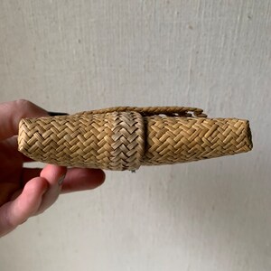 May include: A woven straw clutch bag in a light brown colour. The bag is closed and has a woven flap.