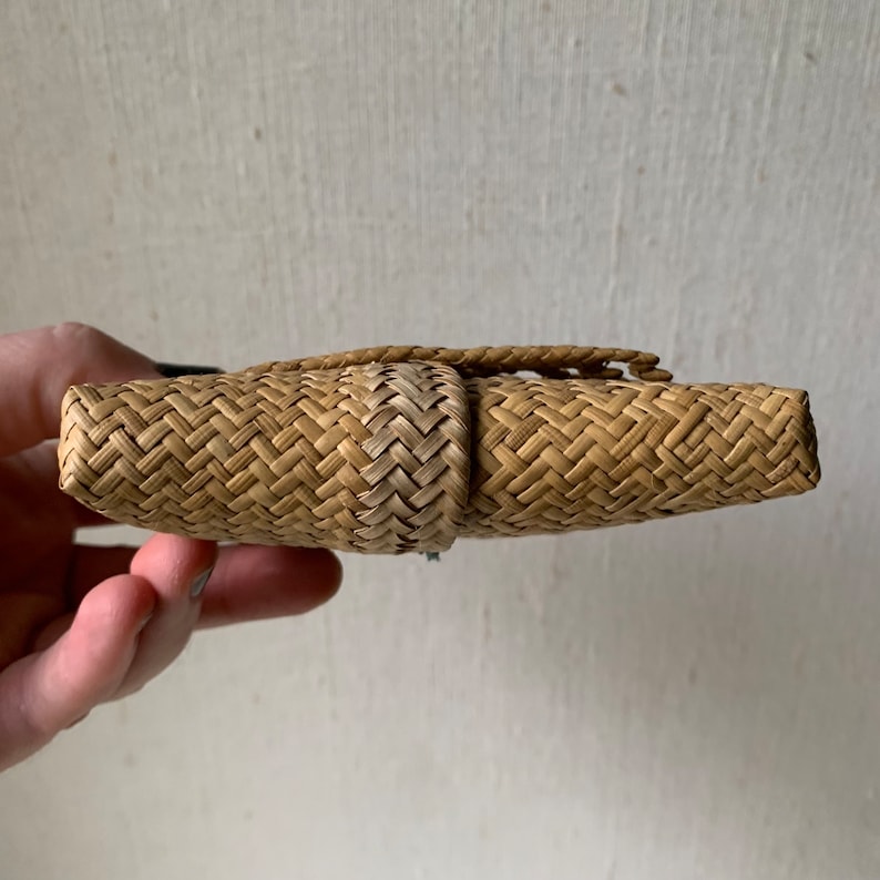 May include: A woven straw clutch bag in a light brown colour. The bag is closed and has a woven flap.