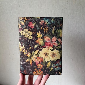 May include: A rectangular journal with a dark background and a colorful floral pattern. The flowers are in shades of yellow, pink, red, and blue. The journal is held in a person's hand.