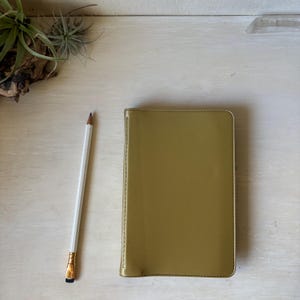 May include: A rectangular notebook with a smooth, metallic gold cover and visible stitching along the spine, positioned next to a white pencil with a gold ferrule and a dark lead tip, providing a clear scale for the notebook's size.