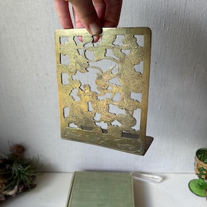May include: A vintage brass bookend with an intricate cut-out design featuring figures, birds, and floral elements. The bookend has a rectangular shape with a small base for stability. The brass has a warm, aged patina, adding to its antique charm.