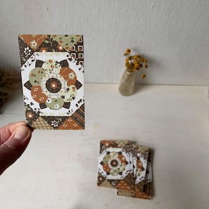 May include: A stack of decorative cards with a floral and geometric pattern in shades of brown, orange, and green. The cards feature a central floral design surrounded by patterned borders. The cards are displayed on a white surface.