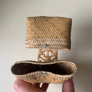 May include: A woven straw basket with a lid. The basket is light brown and has a woven pattern. The lid has a woven pattern and is slightly darker than the basket. The basket is tied together with blue string.