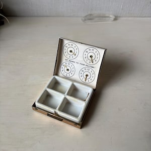 May include: Vintage pillbox with a hinged lid and four compartments. The lid features four clock faces with hands, and the text "SET HANDS TO YOUR PILLTIME!". The box is made of metal with a white interior.