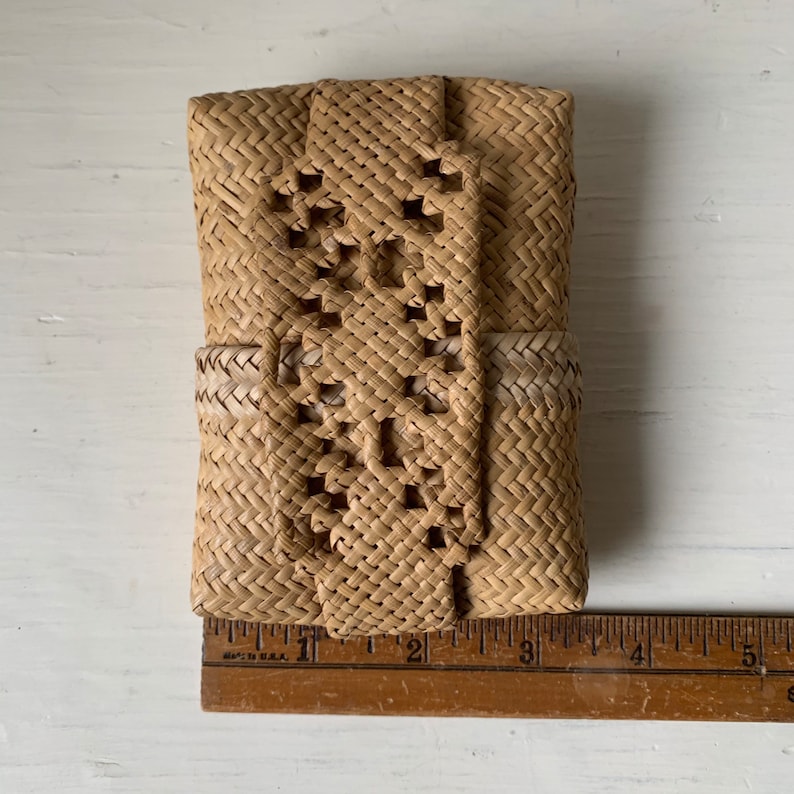 May include: A small, woven, light brown, rectangular bag with a decorative pattern. The bag is approximately 10 cm wide and 15 cm tall.