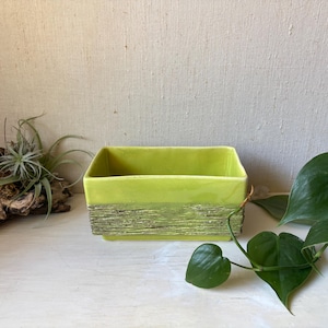 May include: A rectangular ceramic planter with a glossy lime green upper section and a textured, matte olive green base. The vessel features clean, straight lines and a smooth interior finish, providing a structured container for small plants.