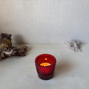 May include: A red glass votive candle holder with a textured, beaded design. The candle holder contains a lit, orange candle. A piece of driftwood and a crystal formation are in the background.