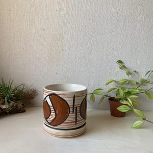 May include: A white ceramic planter with a brown and black geometric design. The planter has a wide base and a slightly flared top.