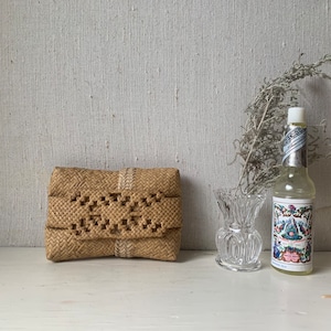 May include: A woven straw clutch bag with a white trim and a decorative pattern. The bag is sitting on a white surface next to a clear glass vase and a bottle of floral water.