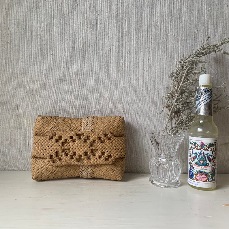 May include: A woven straw clutch bag with a white trim and a decorative pattern. The bag is sitting on a white surface next to a clear glass vase and a bottle of floral water.