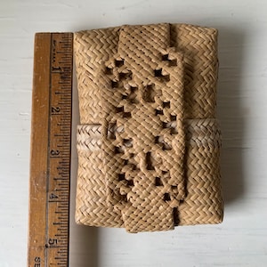 May include: A small, woven, light brown basket with a decorative pattern. The basket is closed with a woven flap.