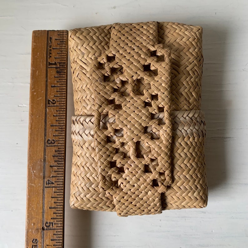 May include: A small, woven, light brown basket with a decorative pattern. The basket is closed with a woven flap.