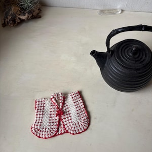 May include: A handmade, red and white crochet tea cozy with a button closure, next to a black cast iron teapot. The cozy has a checkered pattern and is designed to fit around a teapot.