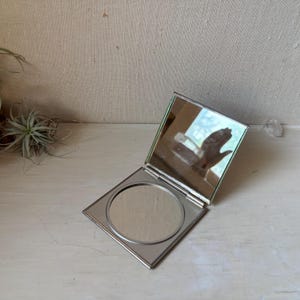 May include: A square, silver-toned metal compact mirror with a hinged lid that opens to reveal a circular mirror surface. The exterior frame is flat and metallic, featuring a clean, reflective finish on both the inner circular glass and the lid.