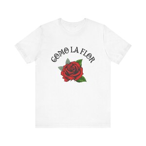 May include: White t-shirt featuring a red rose with green leaves and the text "COMO LA FLOR" in black, arched above the flower. The design is centered on the front of the shirt, creating a simple yet striking visual.