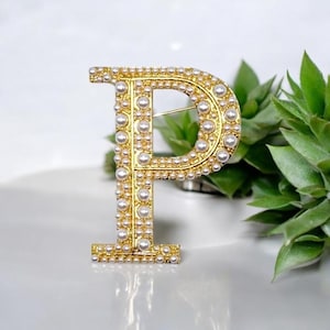 Op de afbeelding: Een gouden broche in de vorm van de letter P, versierd met witte nepparels. De broche ligt op een wit oppervlak met een groene plant op de achtergrond.