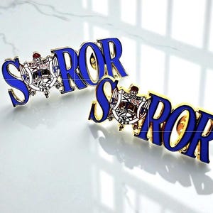 Puede incluir: Dos broches dorados con la palabra "SOROR" en letras de esmalte azul. Cada broche presenta un escudo con las palabras "FOUNDED 1922". Los broches se muestran sobre una superficie blanca.