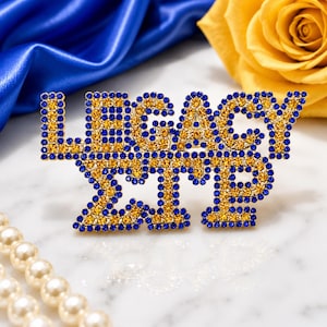 Könnte beinhalten: Eine dekorative Brosche mit dem Schriftzug LEGACY und den griechischen Buchstaben Sigma Gamma Rho, gefertigt mit einer goldfarbenen Metallbasis und besetzt mit kleinen, funkelnden blauen und gelben Strasssteinen in Blockschrift.