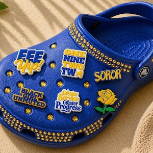 Könnte beinhalten: Ein blauer Clog-Schuh, verziert mit dekorativen Charms und Strasssteinen. Die Charms zeigen Sätze wie "EEE Yip!" und "BLACK GIRL UNMUTED." Eine gelbe Rosen-Grafik ist ebenfalls vorhanden. Der Schuh ist mit goldfarbenen Strasssteinen verziert.