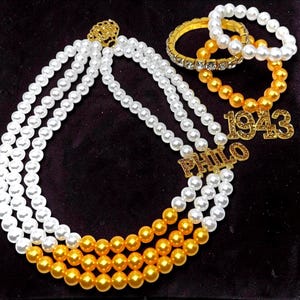 Pode incluir: Um conjunto de colares e pulseiras de pérolas. Os colares apresentam contas brancas e douradas, com pingentes dourados que soletram "PHILO" e o número "1943". As pulseiras são douradas e brancas, com detalhes de strass.