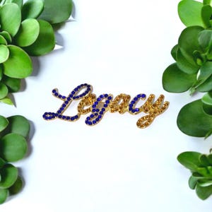 May include: A script-style word pin spelling out the word Legacy. The letters are constructed from a gold-toned metal frame encrusted with alternating blue and amber-coloured rhinestones that catch the light, creating a decorative accessory.