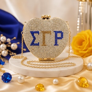 Könnte beinhalten: Eine herzförmige Clutch mit goldenem Glitzer, verziert mit den griechischen Buchstaben Sigma Gamma Rho aus blauen und goldenen Kristallen. Die Tasche verfügt über einen dünnen goldenen Kettenriemen und einen kleinen dekorativen Verschluss.
