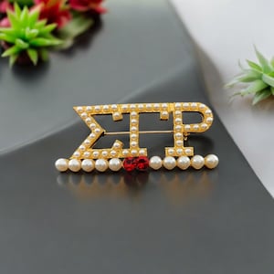 May include: Gold sorority pin featuring the letters "ΣΠΡ" surrounded by white pearls. Two red gems are centred below the letters.
