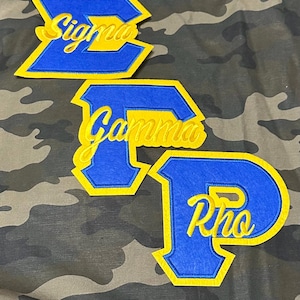 Puede incluir: Tres parches de fieltro azul y amarillo con las letras griegas Sigma, Gamma y Rho en letras amarillas.