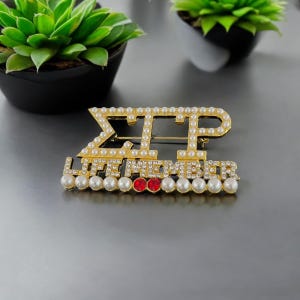 May include: Gold-toned pin with white pearls and red rhinestones. The pin features the letters "ΣΤΡ" and the words "LIFE MEMBER".