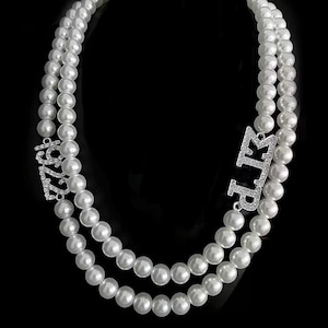 Puede incluir: Un collar de perlas de doble hebra con cierres plateados. Un cierre dice "1922" y el otro "LTP", ambos adornados con pequeñas piedras brillantes. Las perlas son blancas y el fondo es negro.