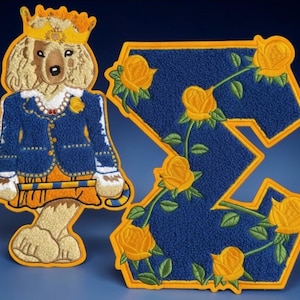 Peut inclure: Écussons brodés. L'un représente un chien royal avec une couronne et une veste bleue à bordures dorées. L'autre est une lettre "Z" ornée de roses jaunes et de feuilles vertes. Les deux ont un aspect texturé, un fond bleu et des bordures dorées. Mesures en cm.