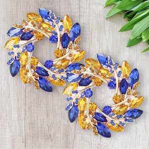 Puede incluir: Dos broches de tono dorado con diamantes de imitación azules y amarillos dispuestos en forma de hoja. Los broches son circulares y tienen un diseño delicado e intrincado.
