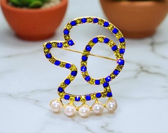 May include: Gold-toned brooch with a stylized design, adorned with blue and yellow gemstones. The brooch features a series of dangling pearls at the bottom. The background includes two small potted plants.