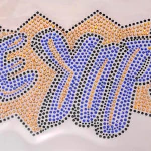 Könnte beinhalten: Nahaufnahme eines weißen Stoffes mit dem Wort "Exxp!" in blauen, schwarzen und goldenen Strasssteinen.