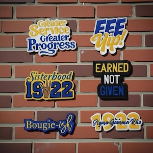 May include: A selection of colourful patches with text displayed on a brick wall. The patches feature phrases such as "Greater Service, Greater Progress," "Sisterhood 1922," and "Earned Not Given." The patches are in various colours, including blue, yellow, and black.