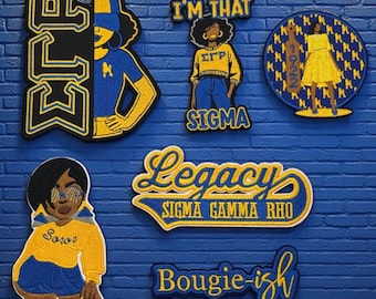 May include: A selection of embroidered patches in blue, yellow, and black. The designs feature sorority symbols, figures, and text including "ELP", "I'M THAT SIGMA", "Legacy SIGMA GAMMA RHO", and "Bougie-ish". The patches are shown against a blue brick wall.