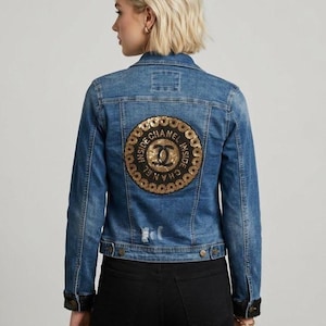 May include: A blue denim jacket featuring a large circular patch on the back with gold and black sequins. The patch displays the text CHANEL INSIDE in a circular pattern around a central interlocking logo. The jacket has long sleeves with black cuffs.