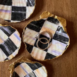 May include: Four decorative ceramic seashell ring dishes with a black and white checkered pattern and gold accents. One dish has two gold rings on top.