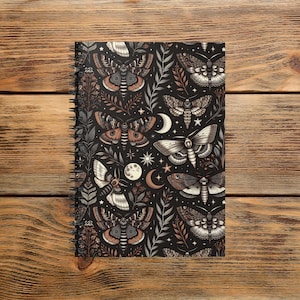 May include: A spiral-bound notebook featuring a dark background with a repeating pattern of moths, moons, stars, and foliage in shades of brown, gray, and white. The notebook has a black spiral binding.