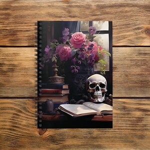 May include: Spiral-bound notebook featuring a gothic still life. The cover art depicts a skull, open book, and a vase of pink roses and purple flowers. A stack of books and a decorative lamp are also visible.