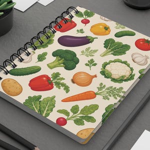 May include: A spiral-bound notebook with a cream-colored cover featuring a repeating pattern of colorful vegetables including broccoli, carrots, red peppers, eggplants, onions, cauliflower, tomatoes, cucumbers, radishes, and garlic cloves.