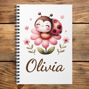 May include: A white spiral-bound notebook featuring a cartoon ladybug resting on a pink flower with green leaves. The design includes small pink flowers and hearts surrounding the central illustration. The name Olivia is printed in brown script below.