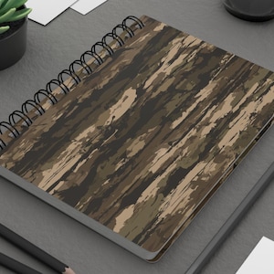 May include: A spiral-bound notebook featuring a camouflage pattern in shades of dark brown, tan, and olive green. The cover has a textured, brushstroke-style print. The notebook is held together by a black metal wire coil along the left side.