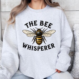 May include: Light gray sweatshirt with a large graphic of a bee and the text "THE BEE WHISPERER" in black. The bee is yellow and black with detailed wings. The sweatshirt is a casual style.