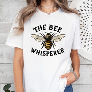 May include: White t-shirt featuring a detailed illustration of a bee with black and yellow stripes, and the text "THE BEE WHISPERER" in black. The bee has tan wings. The shirt is a short-sleeved crew neck.
