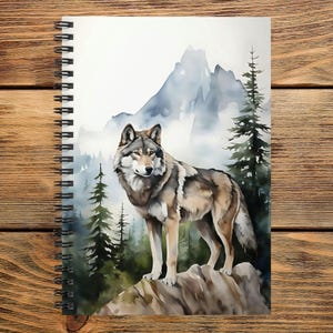 May include: A spiral-bound notebook featuring a watercolor illustration of a gray and brown wolf standing on a rocky outcrop. The background depicts a misty mountain range and tall evergreen pine trees in shades of green, blue, and white.